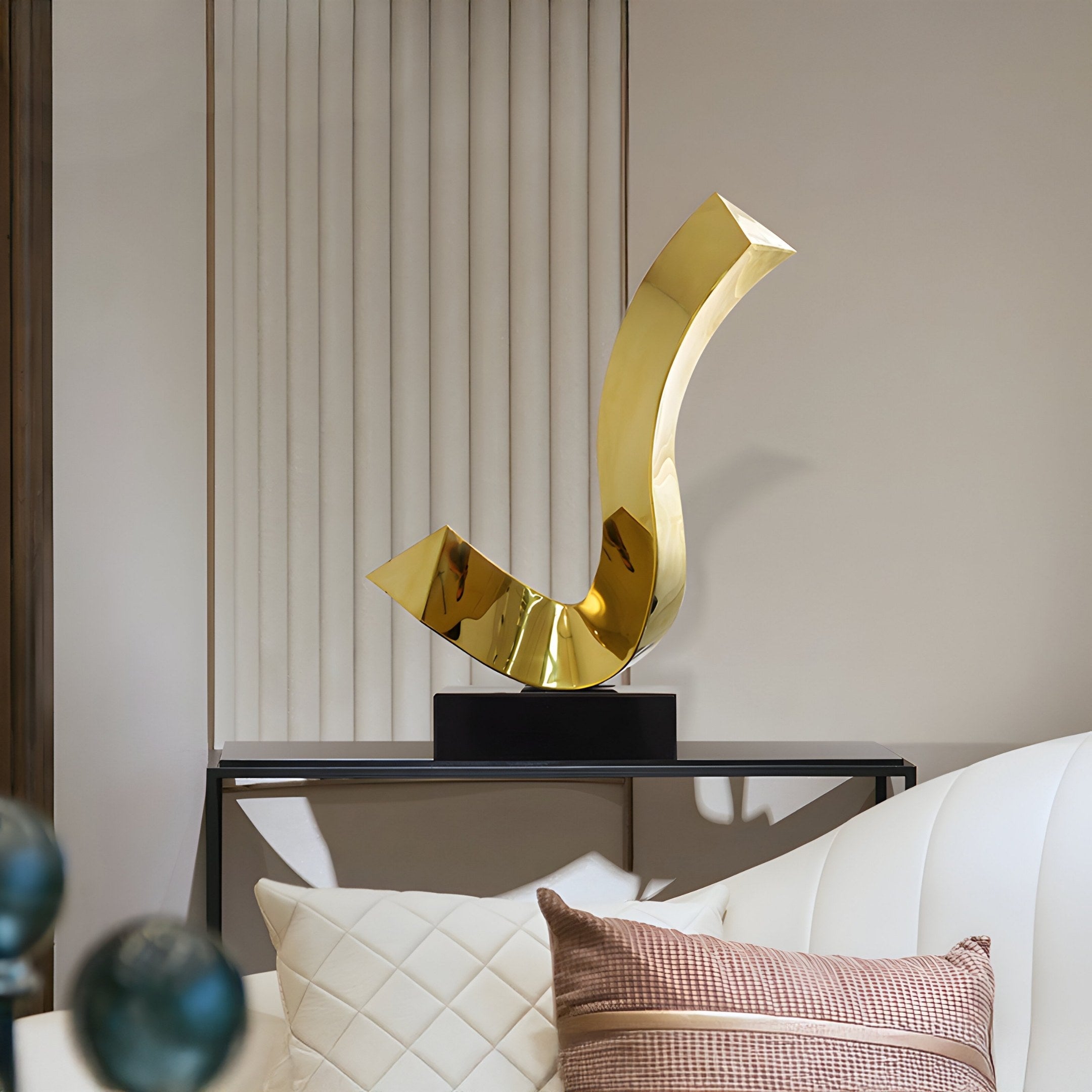 A room presents the Sway Gold Abstract Steel Sculpture by Giant Sculptures on a black pedestal atop a console table against ridged panels. In the foreground, a white sofa with plush cushions pairs beautifully with this modern décor piece.