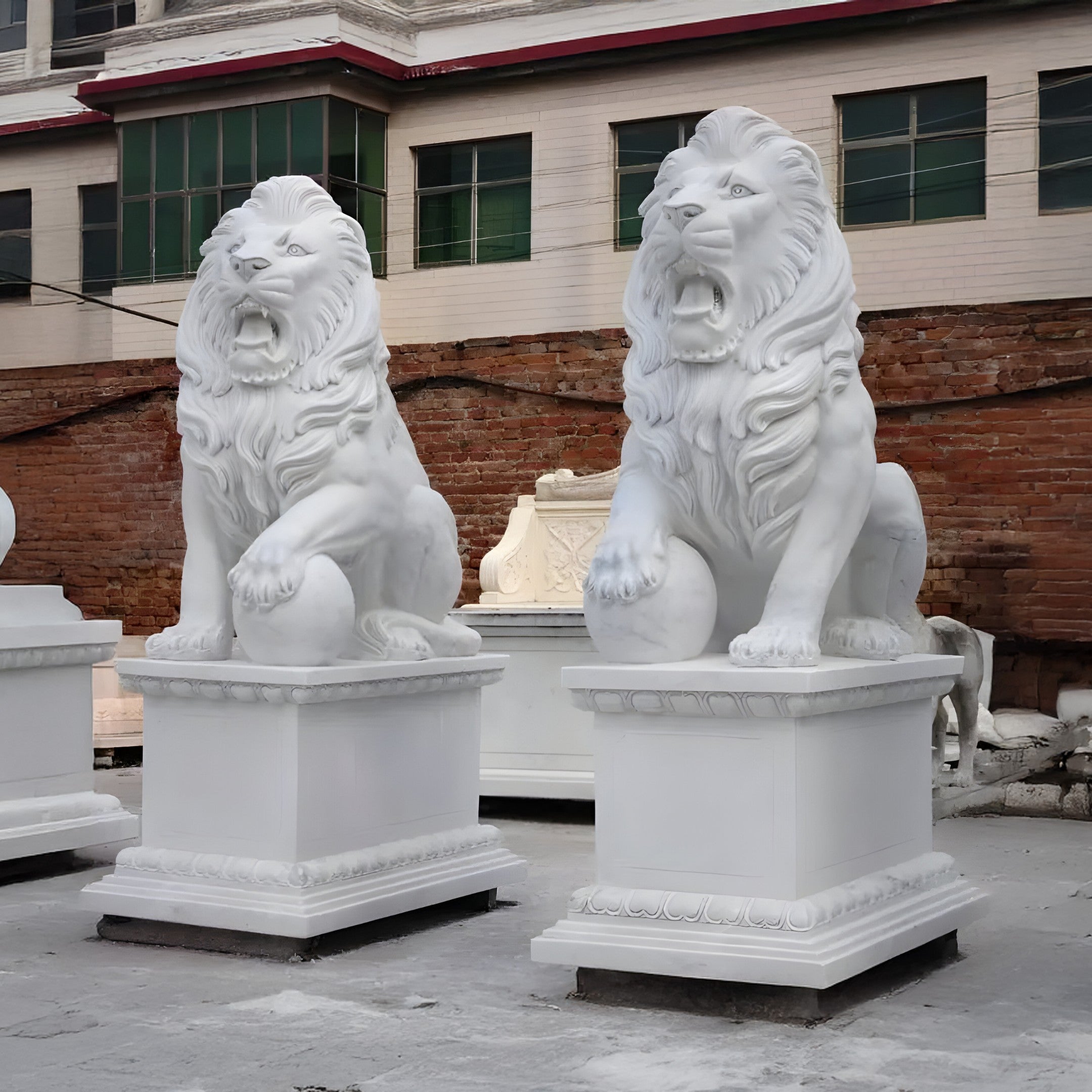 The Giant Lion With Ball Marble Outdoor Sculptures by Giant Sculptures add a majestic touch to the outdoors. Their powerful paws rest on balls atop square pedestals, set against a red brick wall and a beige building with green-framed windows, capturing attention effortlessly.