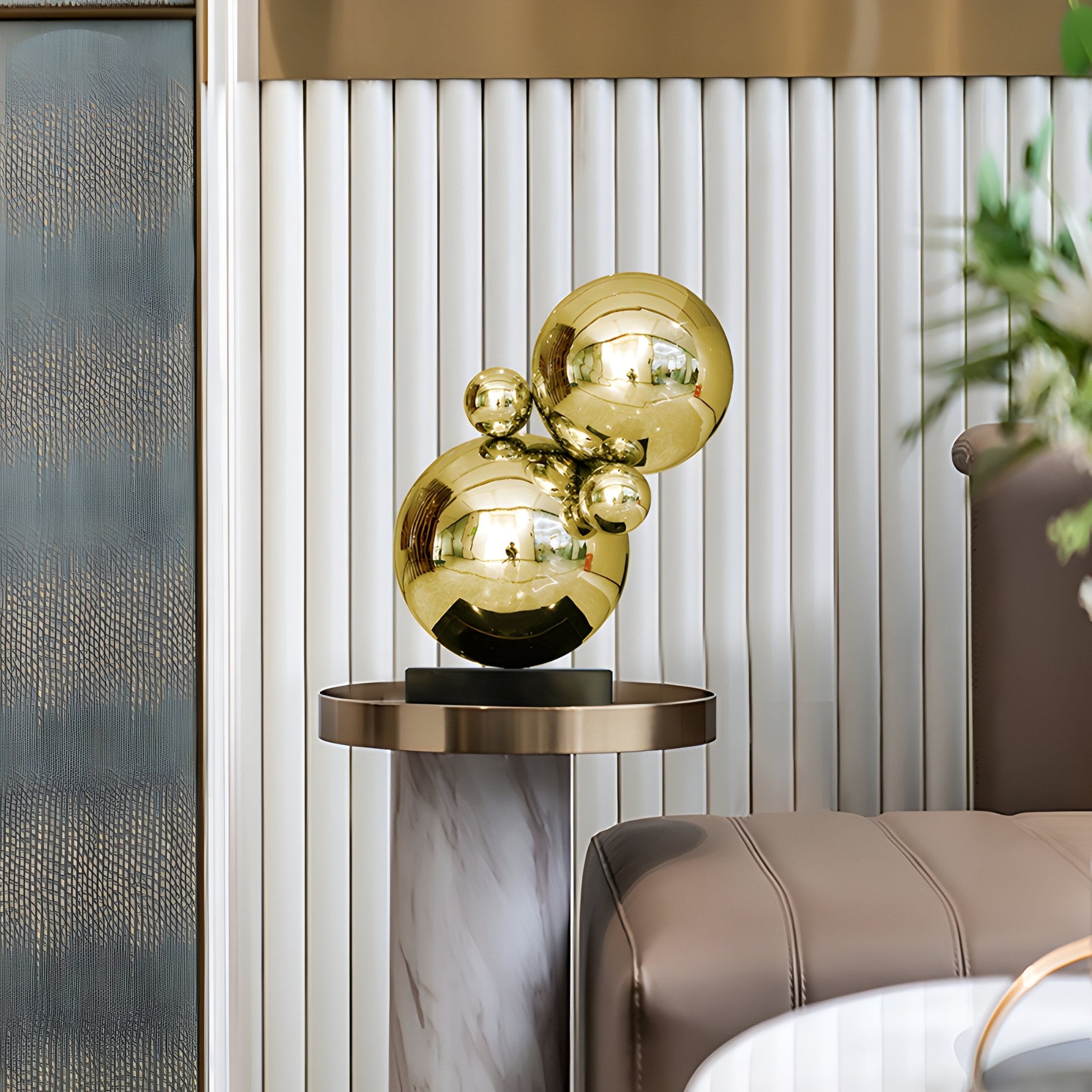 The Molecular II Gold Geometric Steel Sculpture by Giant Sculptures, 55cm, features five interconnected gold spheres on a marble-finished round pedestal. The background has vertical ridged panels, with a brown sofa partially visible to the right.