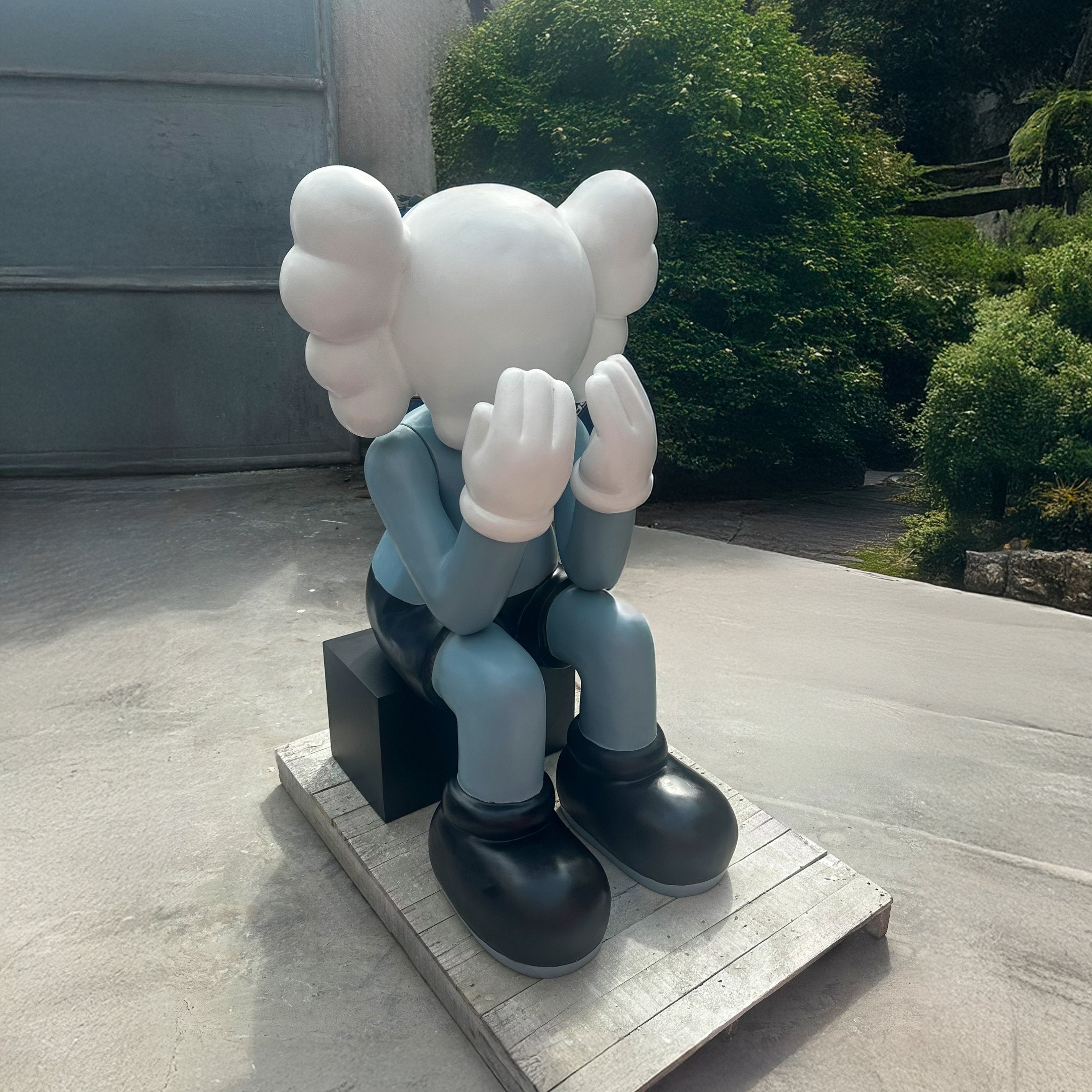 A large outdoor sculpture of a seated character with a white face and round, balloon-like ears, dressed in a blue shirt, black shorts, and large black shoes, hands covering the face in a pose of contemplation or sadness. The sculpture is placed on a wooden pallet, with a lush green background visible behind.