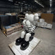 A large indoor sculpture of a seated character with a white face and round, balloon-like ears, dressed in a black-and-white striped shirt, black shorts, and large black shoes, hands covering the face in a pose of contemplation or sadness. The sculpture is positioned on a wooden pallet in an industrial setting, surrounded by warehouse items and boxes.