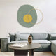 The modern living room, with calming tones, showcases the Solaro Calming Green & Yellow Round Mural Wall Art by Giant Sculptures featuring a large circle and two intersecting rings. It includes a gray sectional sofa, a round coffee table with a textured vase and book, and light wood flooring.