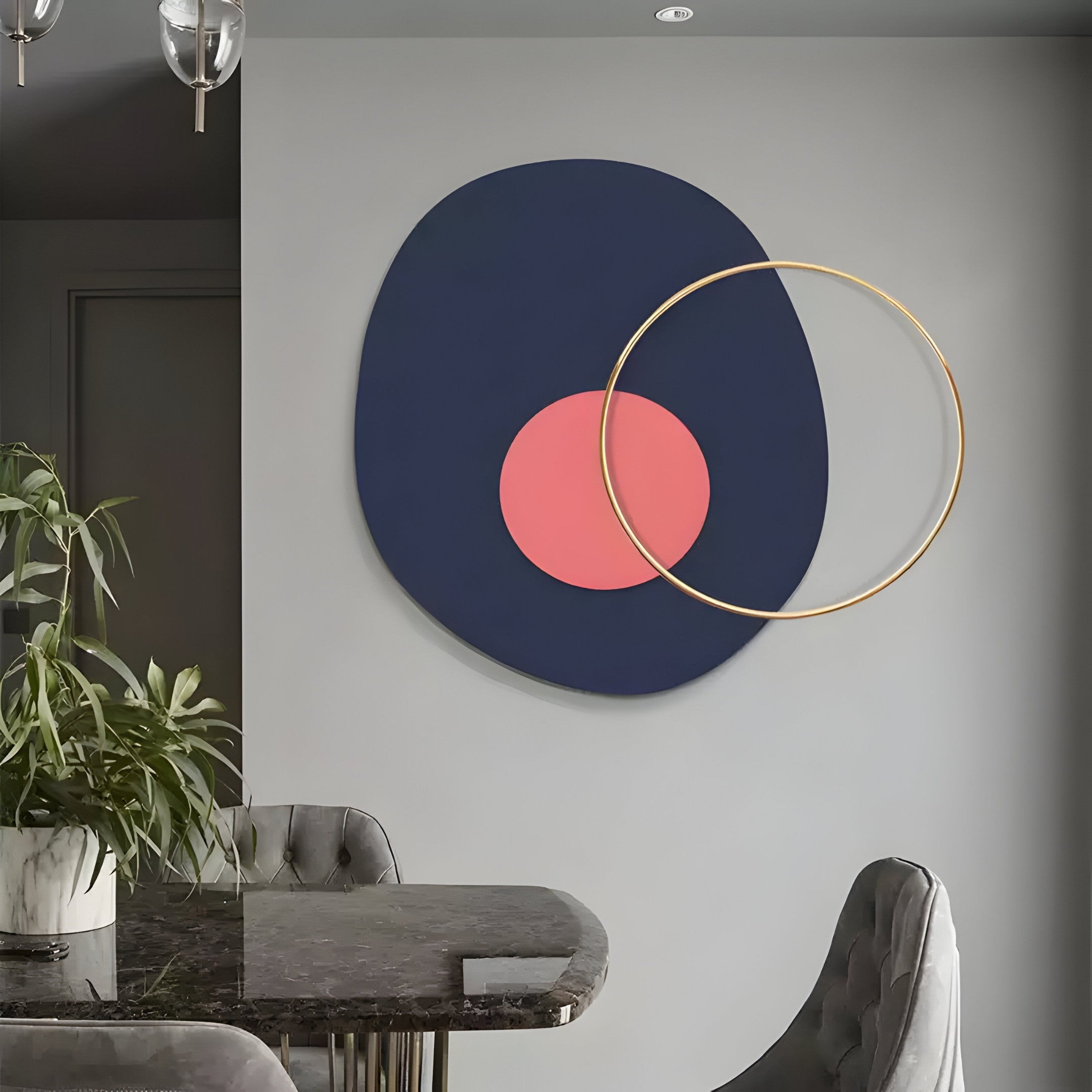 A modern dining room boasts a marble table, grey velvet chairs, and a potted plant. The Solaro Bold Navy & Coral Round Mural Wall Art by Giant Sculptures features abstract shapes with a gold ring on a grey wall. Glass-covered ceiling lights enhance the sophisticated ambiance.