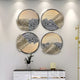 The Aurelio Bottom-Right Metallic Layered Textured Round Wall Art by Giant Sculptures showcases four circular pieces with abstract black, beige, and gray patterns. Subtle gold and silver tones complement their display above a sleek black and white cabinet with a lamp and vase.