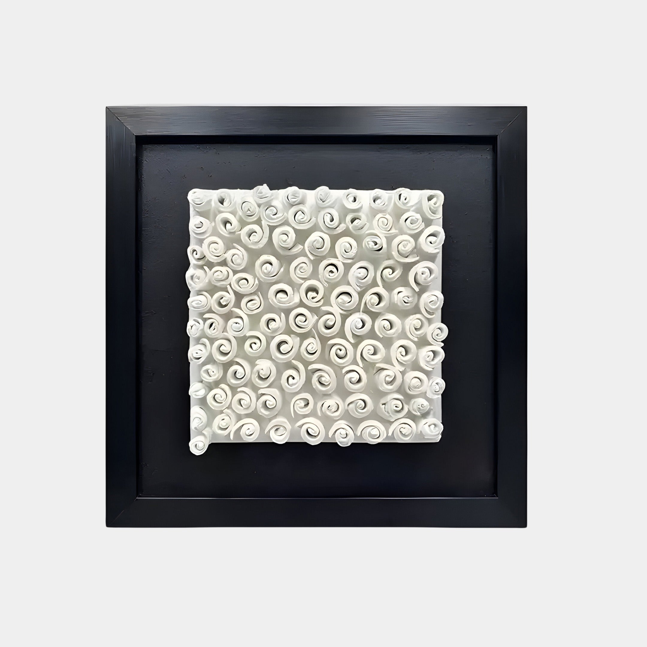 A white 3D artwork featuring an intricate array of rolled, spiral-like shapes resembling scrolls or organic swirls. The sculpture is framed in a black square frame against a dark background.