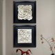 Two black-framed pieces of wall art stacked vertically. The top frame contains a white sculptural rose, and the bottom frame showcases flowing, wave-like white textures.