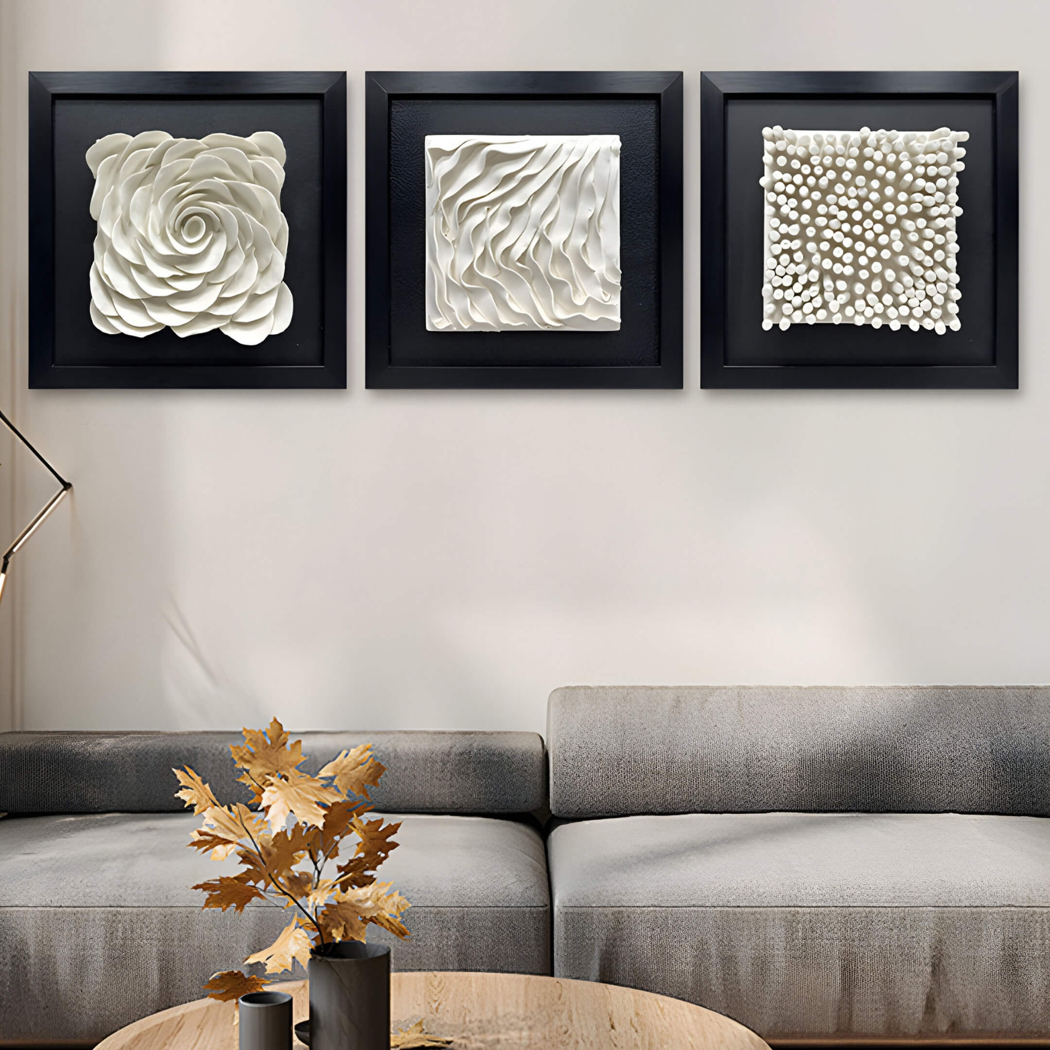 A living room wall with three black-framed wall art pieces arranged horizontally. The left frame has a white sculptural rose, the middle frame displays wave-like white patterns, and the right frame features cylindrical, textured white protrusions.