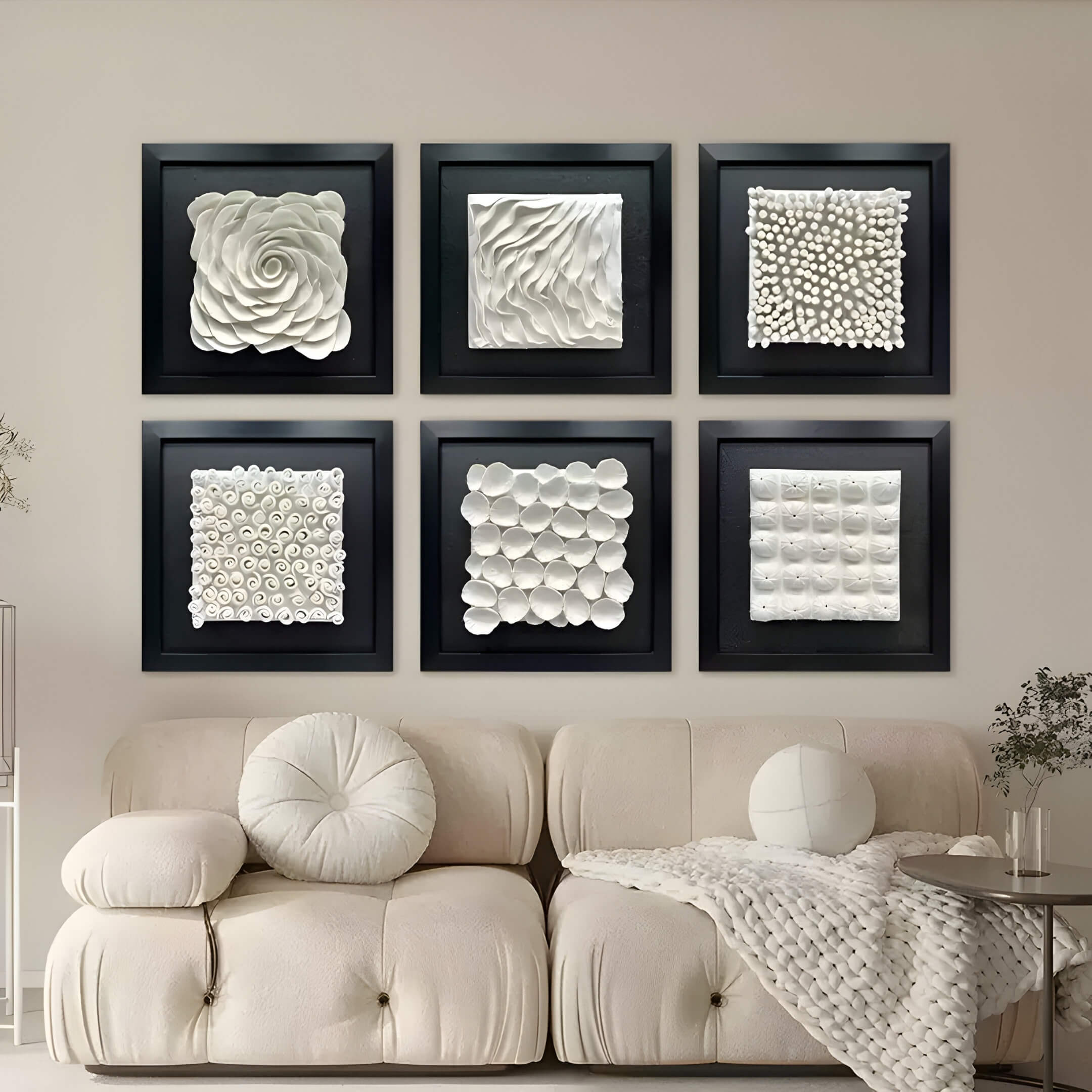 A beige sofa with round and square cushions rests in front of a wall featuring six framed Harmonia 3D Spiral Wall Sculptures by Giant Sculptures, each showcasing unique white flower patterns on a dark background. A knitted blanket and small plant add texture to this modern decor scene.