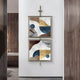 The Vortice Abstract Geometry Dual Panel Wall Art by Giant Sculptures, displaying bold shapes in blue, gold, brown, and white, is framed in gold and mounted vertically on a minimalist system in a modern hallway with wooden paneling.