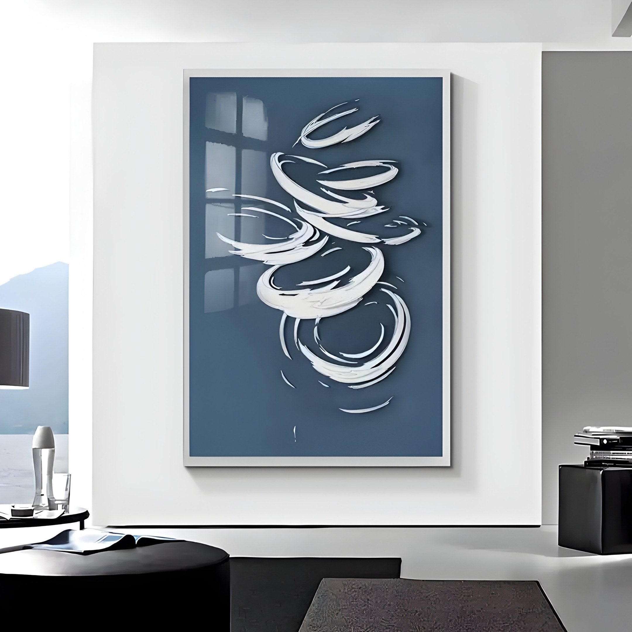 In a modern living room, the Zephyr Azure Swirl Abstract Metal Wall Art by Giant Sculptures takes center stage. Featuring swirling white shapes on blue, it harmonizes with a monochromatic scheme, sleek furniture like a table and books, and expansive windows showcasing a mountain view.