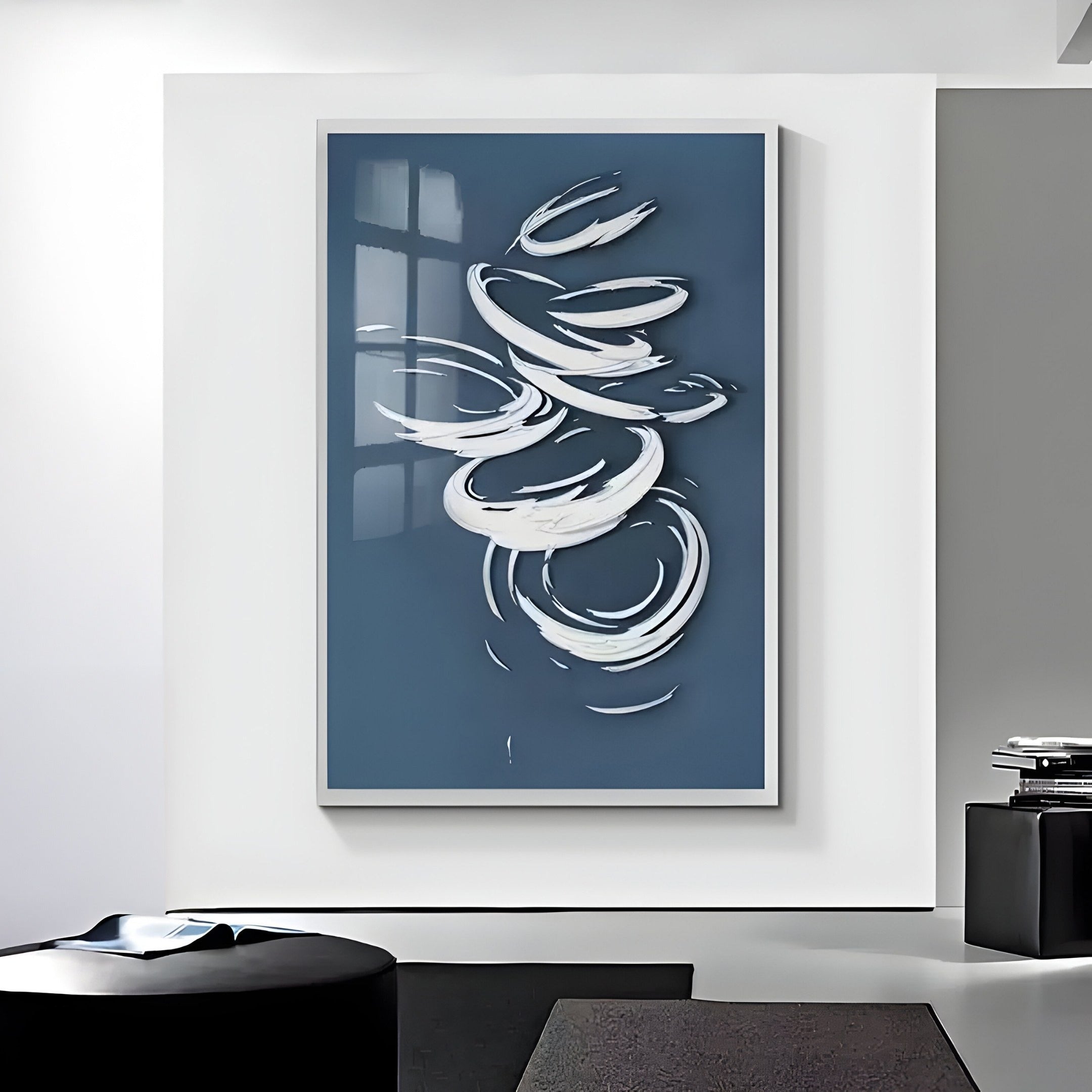 The minimalist decor of a room is enhanced by Giant Sculptures Zephyr Azure Swirl Abstract Metal Wall Art, featuring swirling white designs on a dark blue background. Displayed on a white wall with a black chair and side table with small stacked books, it offers a contemporary visual experience.