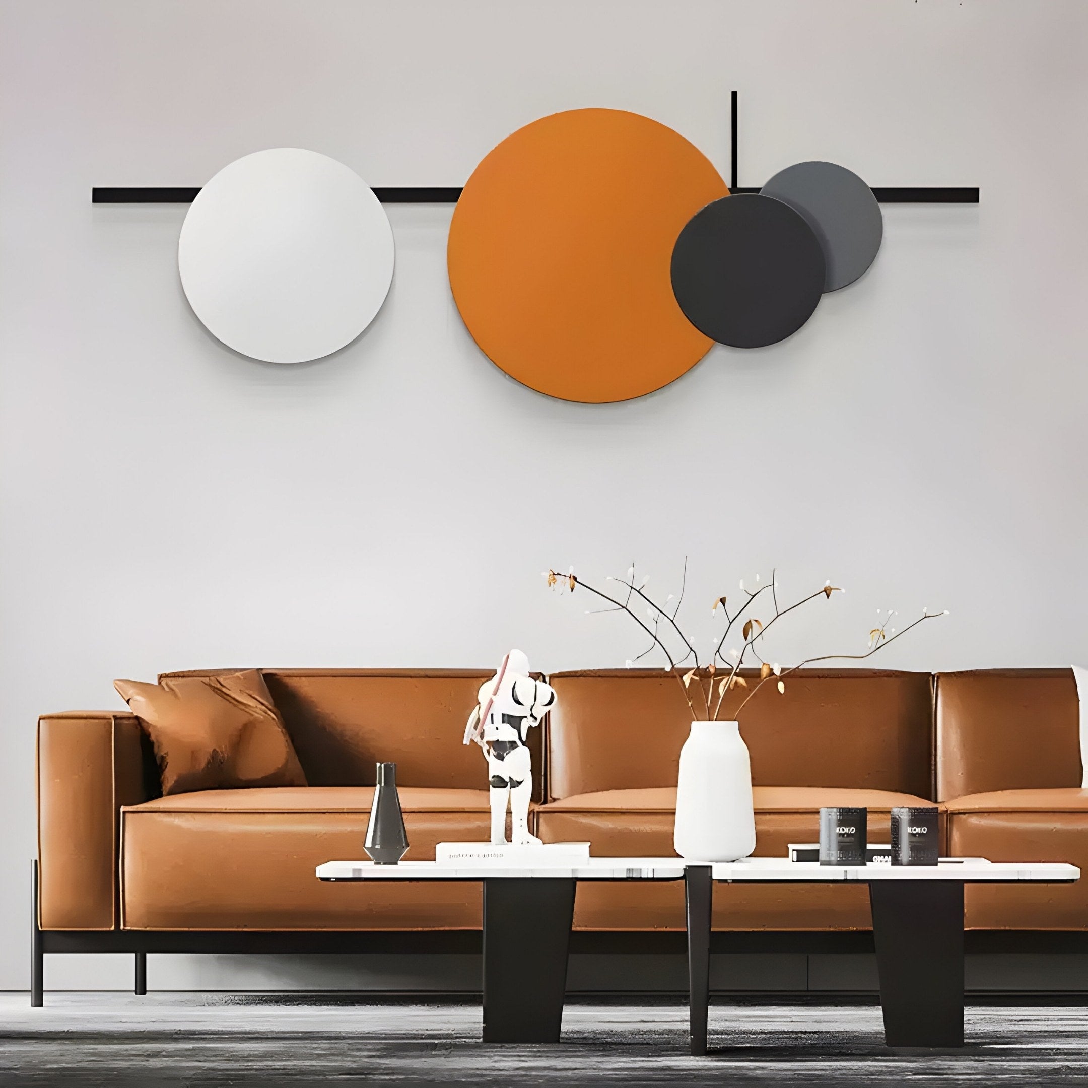 A modern living room showcases a brown leather sofa, a striking black and white figurine, and a vase on a black coffee table. The space is enlivened by Giant Sculptures Orbit II Dynamic Flow Multicoloured Circular Wall Art, adding vibrant colors with its overlapping circles in white, orange, and gray behind the sofa.