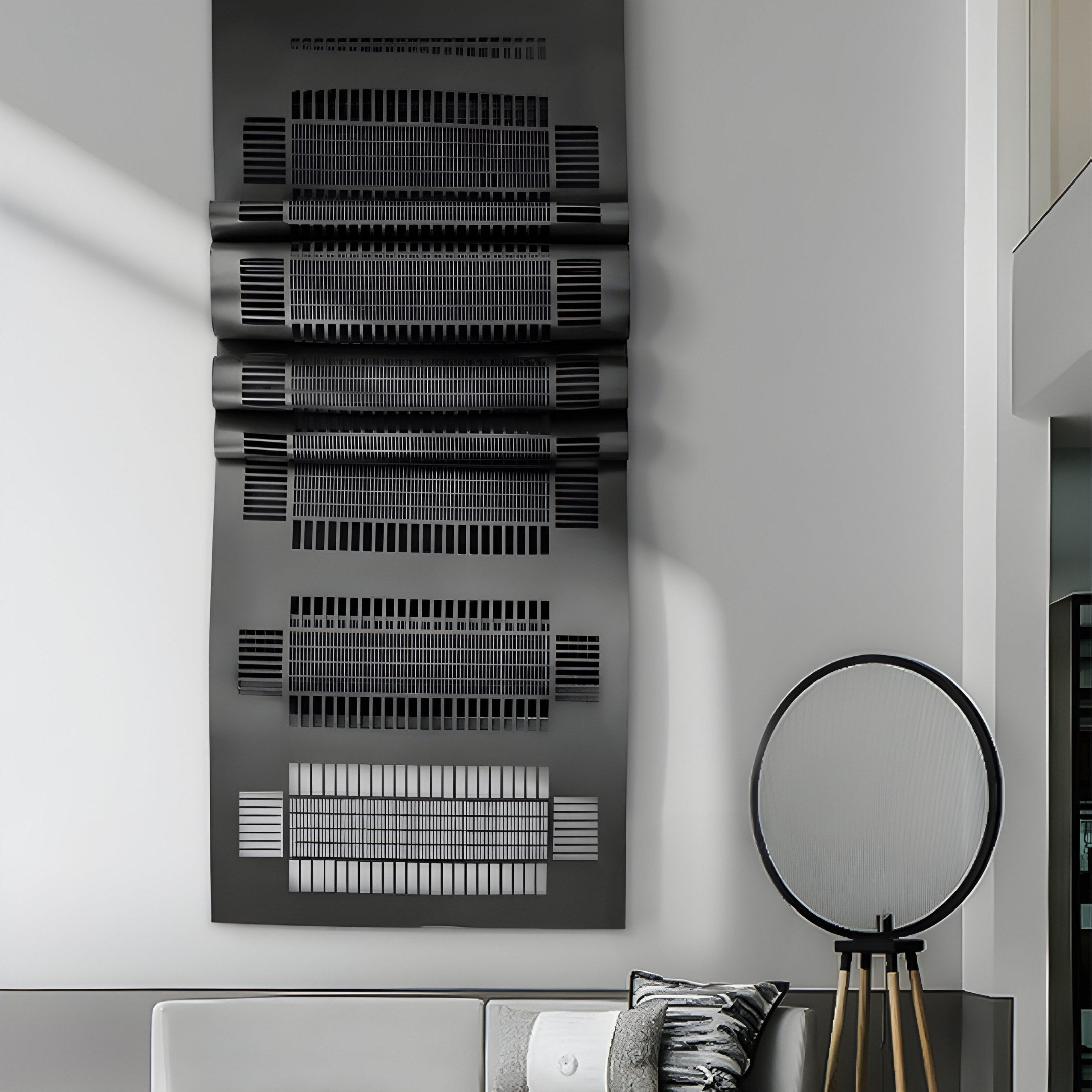 A modern interior showcases the Strata Black Vertical Folded Grid Metal Wall Art by Giant Sculptures, featuring bold geometric patterns. Nearby stands a tall, sleek fan with a circular head, and at the bottom, a gray sofa adorned with patterned cushions is visible.