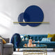 A modern living room boasts a blue sofa, colorful pillows, and a glass coffee table with drinks and a tray. Circular gray abstract art adorns the wall alongside Giant Sculptures Luna Forma Midnight Blue Round Brass Accent Wall Art, highlighted by a slim horizontal light fixture.