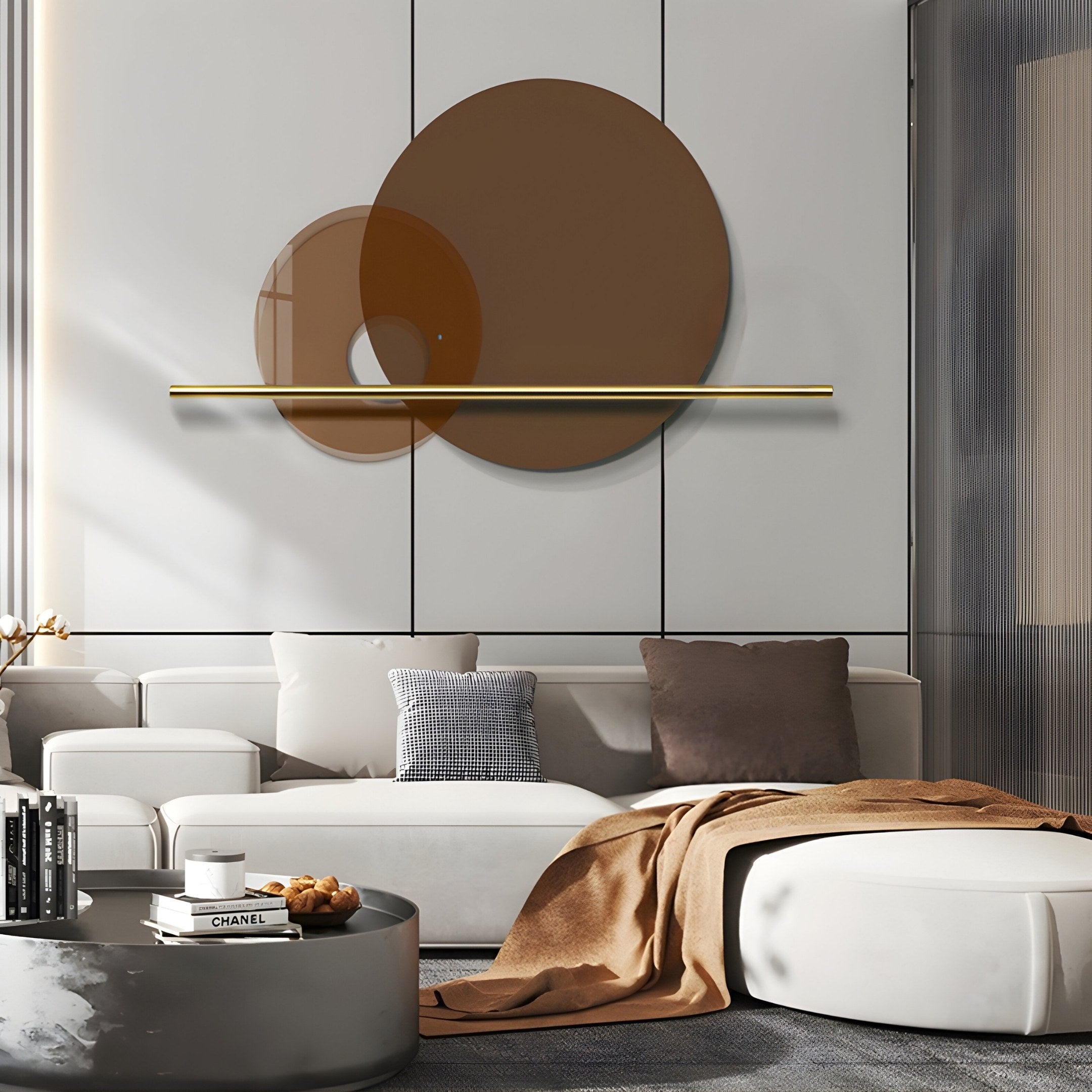 A modern living room features a white sofa with assorted cushions, accented by a round ottoman and beige throw blanket. Highlighting geometric minimalism, two intersecting pieces of the Luna Forma Mocha Brown Round Brass Accent Wall Art by Giant Sculptures add a sleek touch to the space.