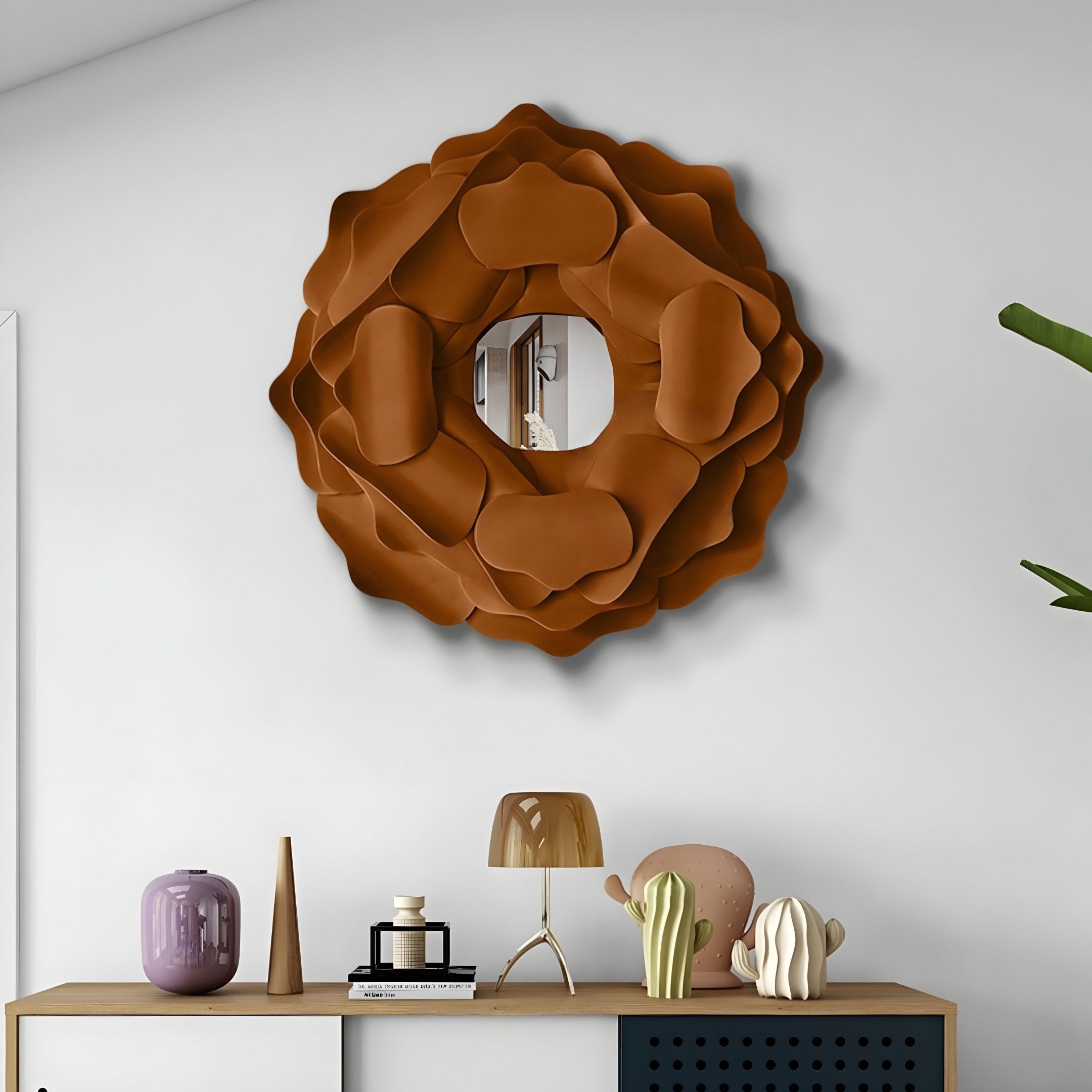 A stylish room showcases the Sculptura Mirror Warm Brown Floral Round Metal Wall Art by Giant Sculptures. Beneath it, a modern console table displays decor like a purple vase and metal sculpture, complemented by a wooden lamp, plant sculptures, and a green plant visible on the right.