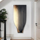 The Celestial Luminous Skyline Curved Vertical Abstract Metal Wall Art by Giant Sculptures, with its modern trapezoidal shape, hangs in the hallway. It features a black base with gold vertical lines and gradient effects, exuding contemporary sophistication alongside a nearby potted plant and staircase.