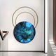 A modern art installation by Giant Sculptures features the Eclipse Luxe Azure Radiance Circular Ring Wall Art: a large, round azure painting with eye-like textures, encircled by overlapping golden rings. Mounted on a white wall in a minimalist space, it embodies contemporary interior elegance.