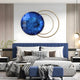 A modern bedroom has a blue upholstered bed with checkered and solid pillows, matched by a houndstooth throw. A Giant Sculptures Eclipse Luxe Sapphire Blue Circular Ring Wall Art and gold rings adorn the wall. Pendant lights hang from the ceiling, and plants on the side table enhance tranquility.