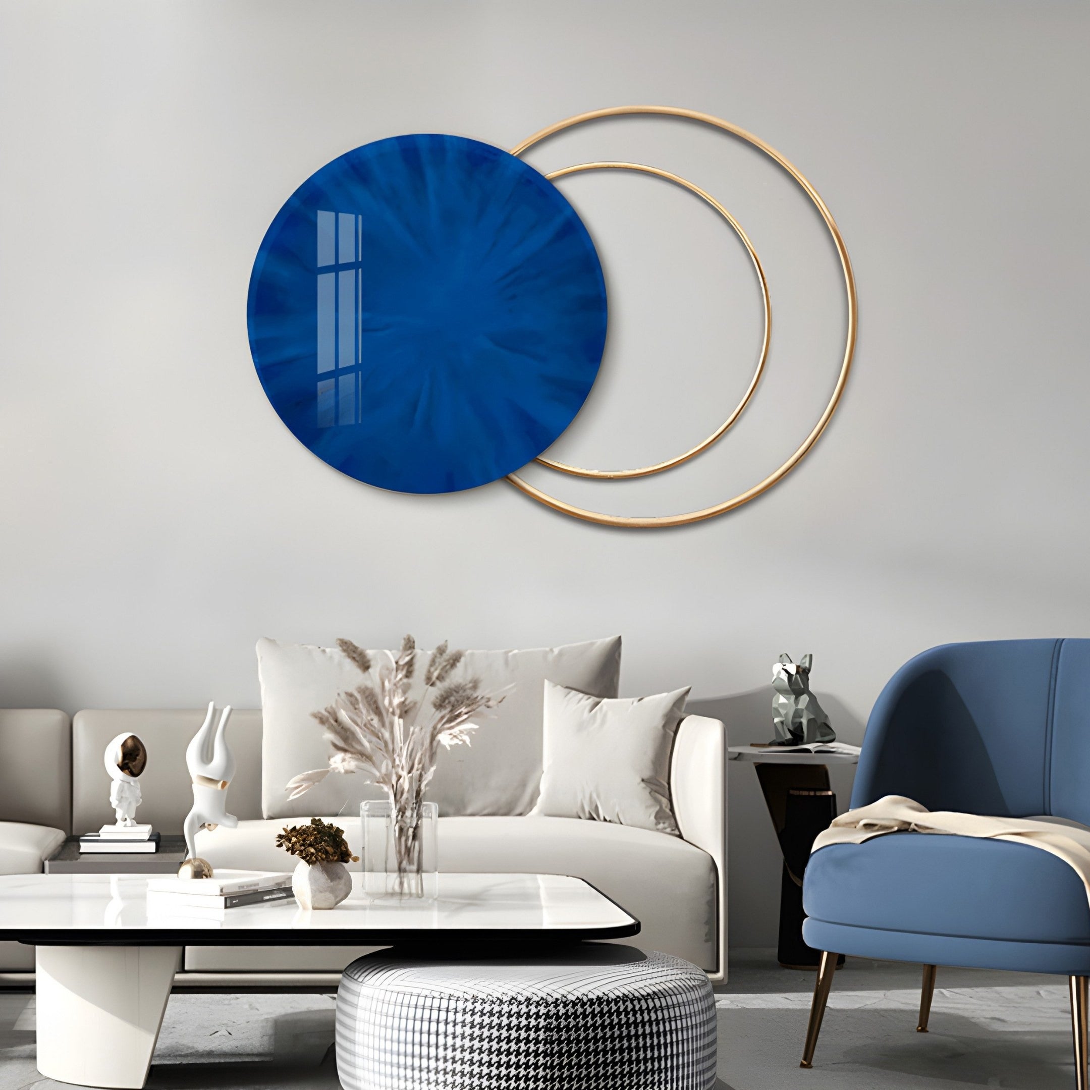 A modern living room features a white sofa, cushions, a white coffee table with a vase of dried flowers, and a blue armchair. The space is enhanced by Giant Sculptures Eclipse Luxe Midnight Blue Circular Ring wall art against the light gray wall.