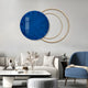 A modern living room features the Eclipse Luxe Midnight Blue Circular Ring Wall Art by Giant Sculptures, with its two gold overlapping rings. It includes a white sofa with beige cushions, a round white coffee table, a blue armchair, and a side table adorned with elegant decor.