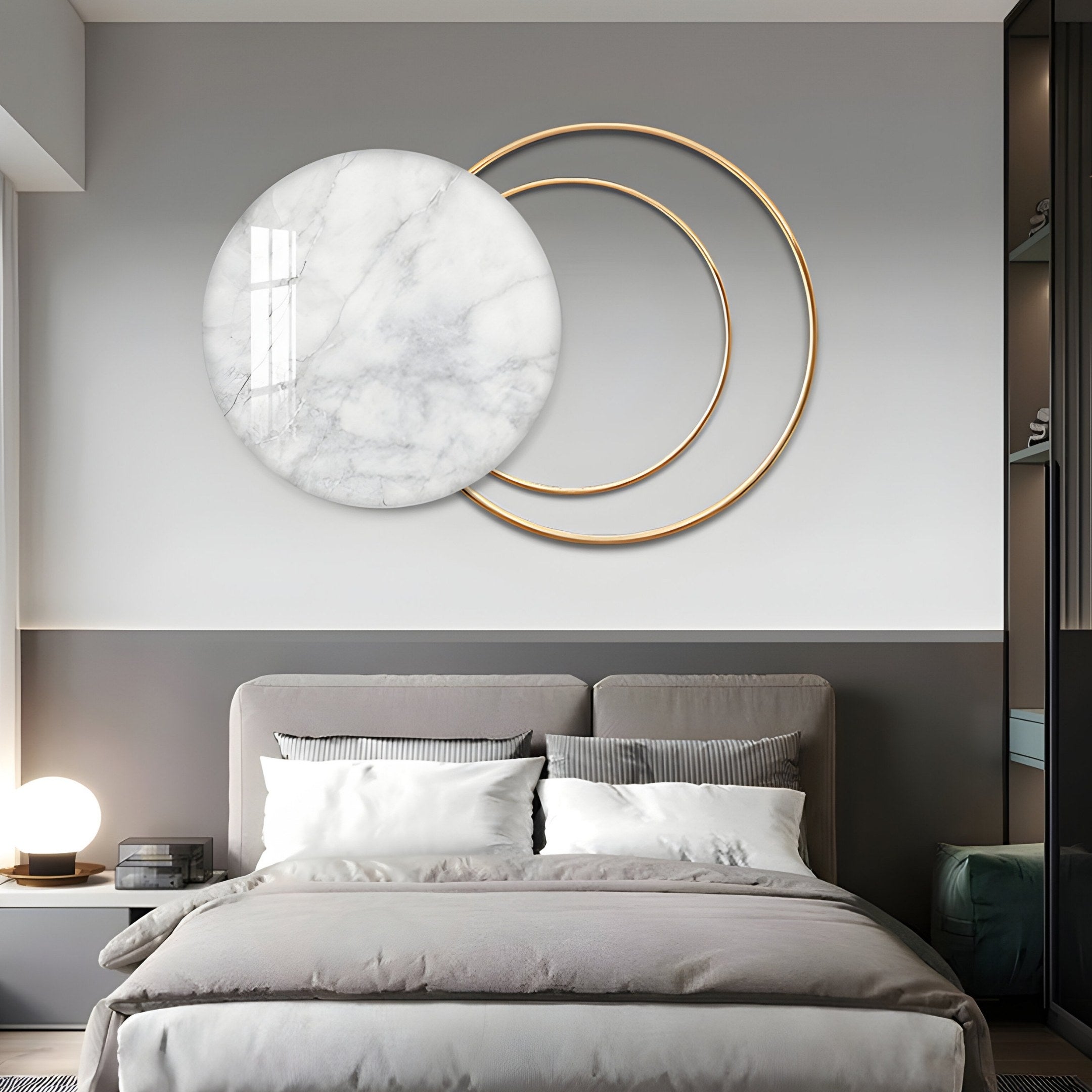 A modern bedroom embodies minimalist charm with its gray and white palette. The headboard matches the bedding, while above, the Eclipse Luxe Bianco White Marble Circular Ring Wall Art by Giant Sculptures displays two overlapping gold rings. A round lamp on the bedside table enhances the design.