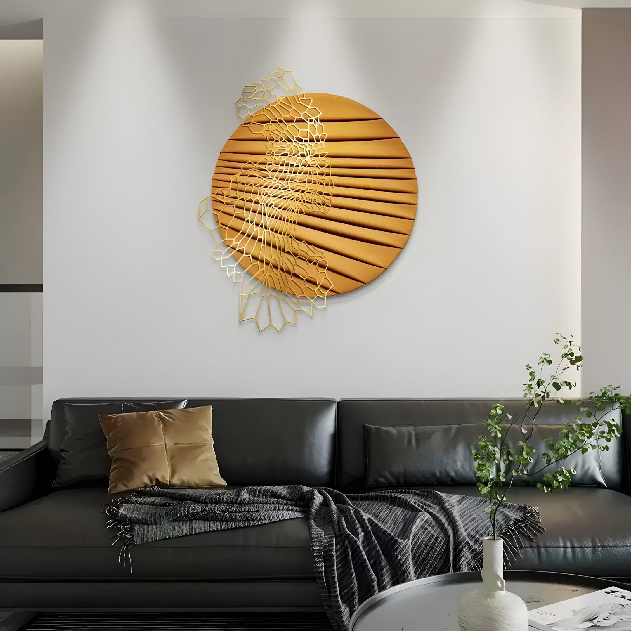 In a modern living room, a dark leather couch is adorned with a brown cushion and gray throw blanket. A light vase with green branches sits on a table nearby, while the white wall features Giant Sculptures Auramesh Sunset Orange-II Metal Winged Circular Geometric Wall Art.