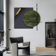 A modern living room with a gray armchair and black floor lamp. The wall features the Tesselle Black Metal Fold & Green Textured Geometric Wall Art by Giant Sculptures, showcasing a black square, gold-curled corner, and green circle intersected by lines. A minimalist shelf with vases completes the design.