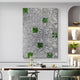 A contemporary dining room features a large Prismatica Green & Silver Mosaic 3D Wall Art from Giant Sculptures. A dining table, decorated with books and items, is surrounded by stylish chairs. Built-in shelves display potted plants and elegant decor, completing the sophisticated look.