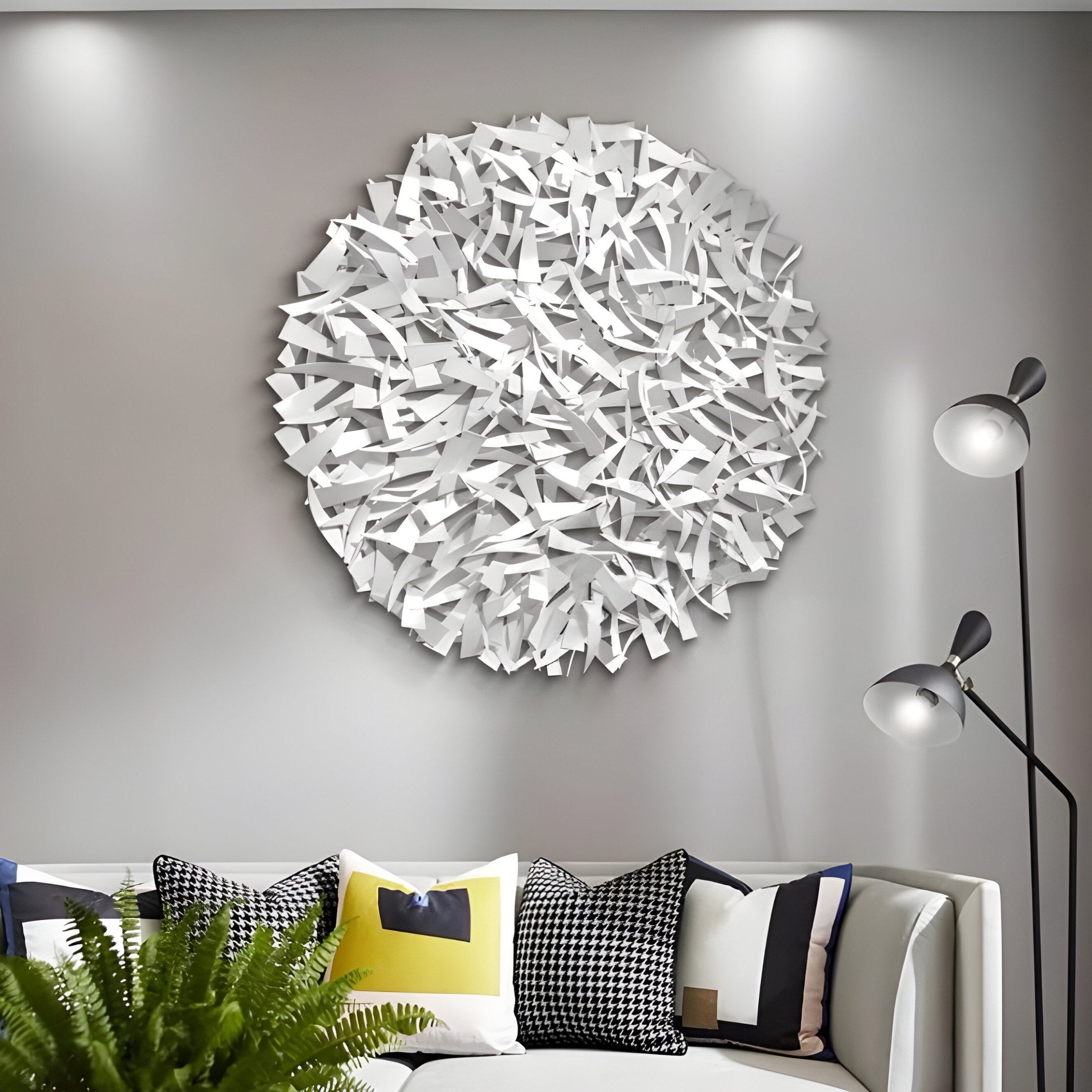 A modern living room features the Eclat Sphere White Abstract Metal Wall Art by Giant Sculptures on a gray wall. Below, a white couch holds black, white, and yellow geometric-patterned pillows. A sleek floor lamp stands nearby, complemented by a potted fern for natural elegance.