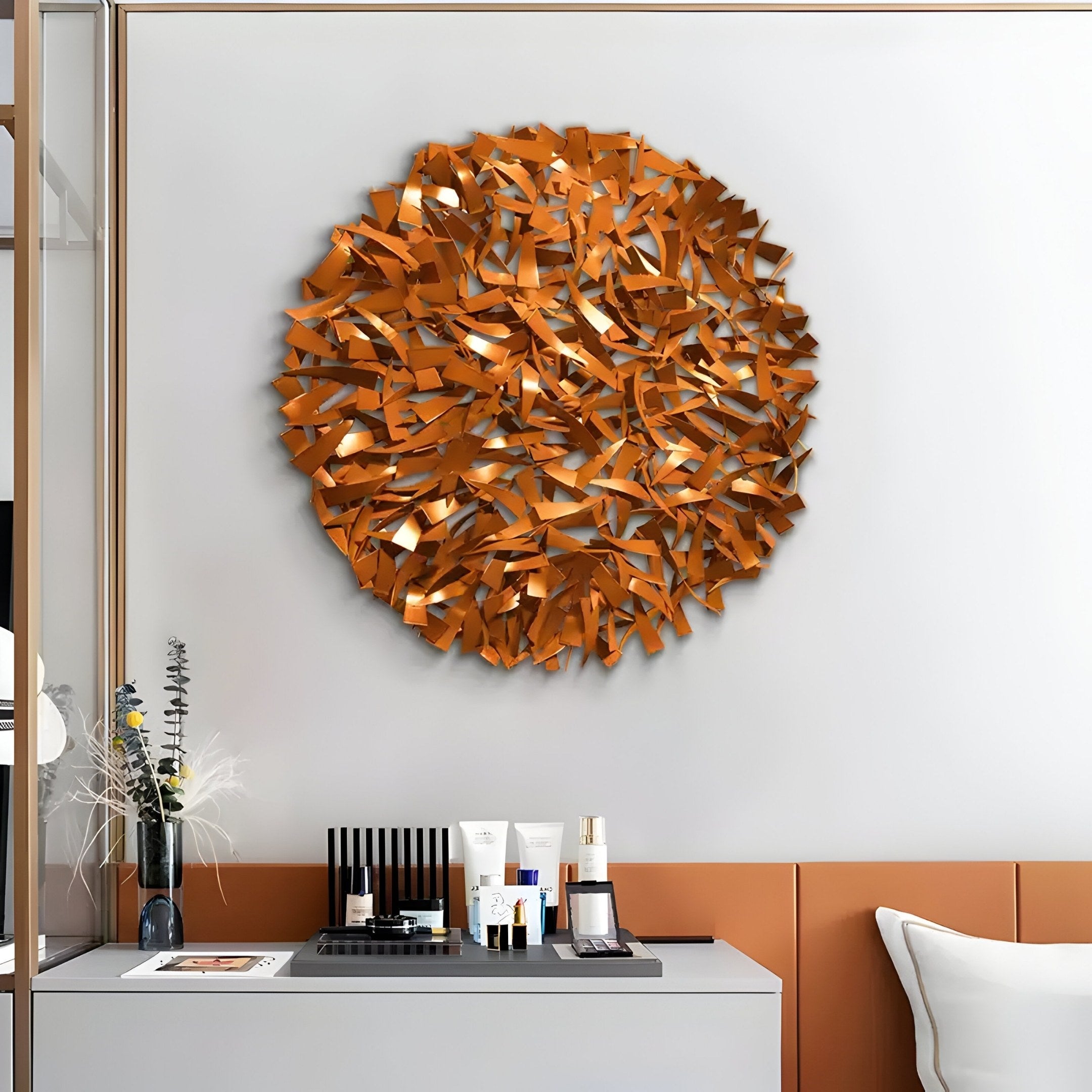 A modern living room features the Eclat Sphere Copper Abstract Metal Wall Art by Giant Sculptures, infusing sculptural design in vibrant orange. Below it, a console table holds decorative items like a vase with dried flowers, black and white boxes, and candles, highlighting the neutral and orange color scheme.