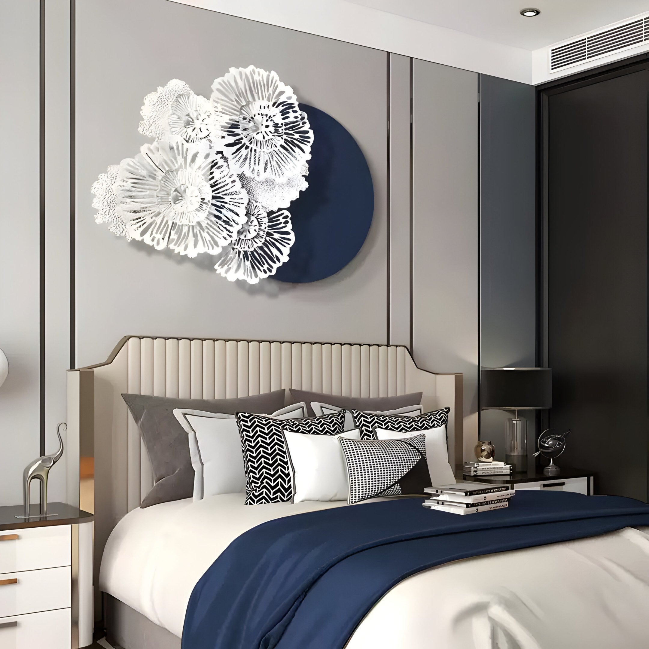 A modern bedroom showcases a stylish bed adorned with white and navy bedding, decorative pillows, and is complemented by the Fleur Halo Navy Blue Round White Metal Floral Decorative Wall Art by Giant Sculptures. A bedside table with books and a lamp completes the sophisticated decor.