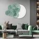 A modern living room with green sofas, patterned cushions, a round coffee table, and decor exudes floral charm. The Fleur Halo White Round Green Metal Floral Decorative Wall Art by Giant Sculptures adorns the space, while shelves filled with books and decor enhance its elegance.