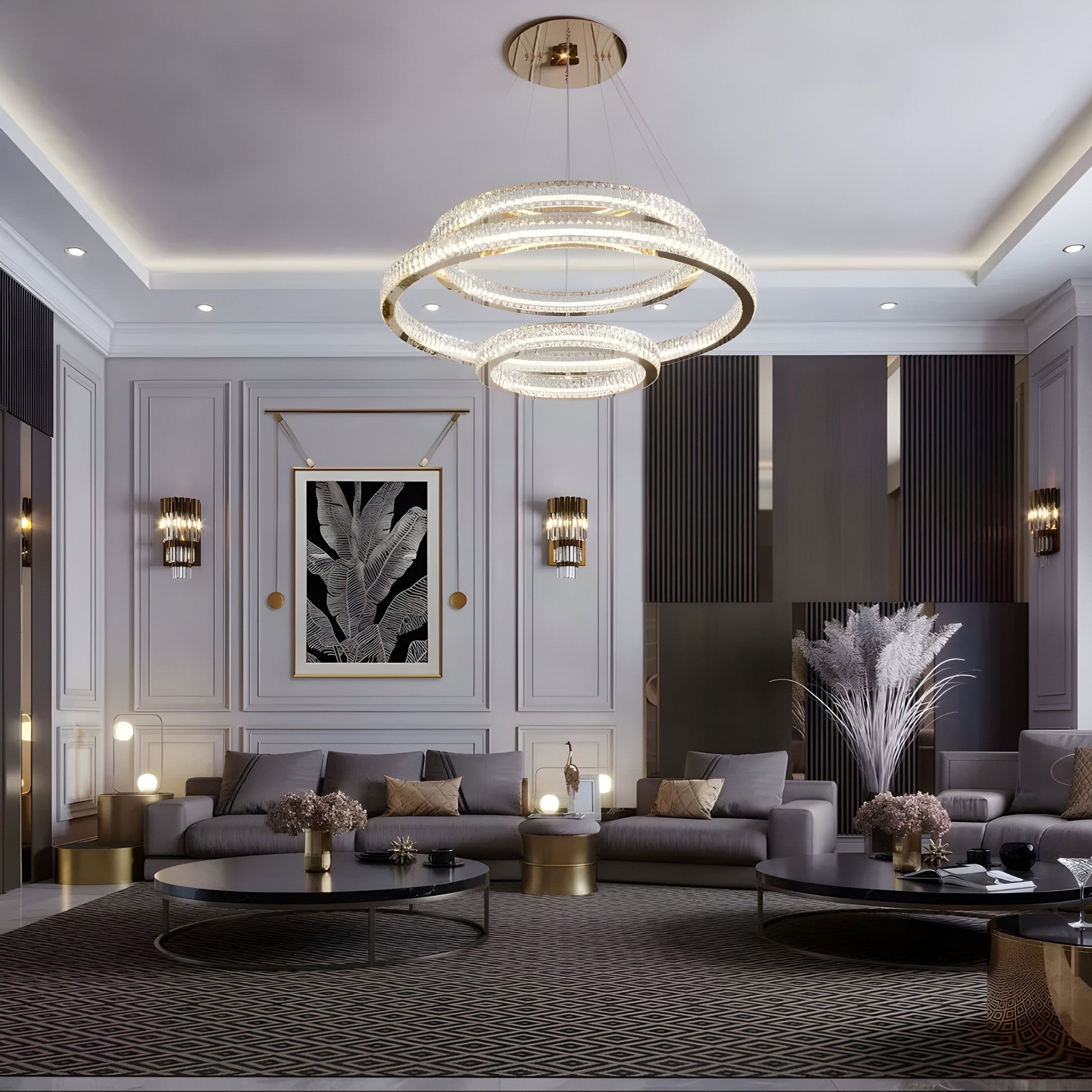 A luxurious living room showcases modern decor with monochrome elegance, featuring two circular chandeliers, gray sofas, and gold-accented coffee tables. The Glacia Monochrome Botanical Crystal Hanging Framed Wall Art by Giant Sculptures adds charm near pampas grass by the sectional couch in tastefully paneled walls.