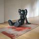 In an art gallery, the Obsidian Black Iconify Daydream Sculpture by Giant Sculptures, measuring 85cm, rests on a textured red and beige rug. With round ears and an expressionless face, it reclines effortlessly on its hands with legs extended across the wooden floor, reflecting modern relaxation.