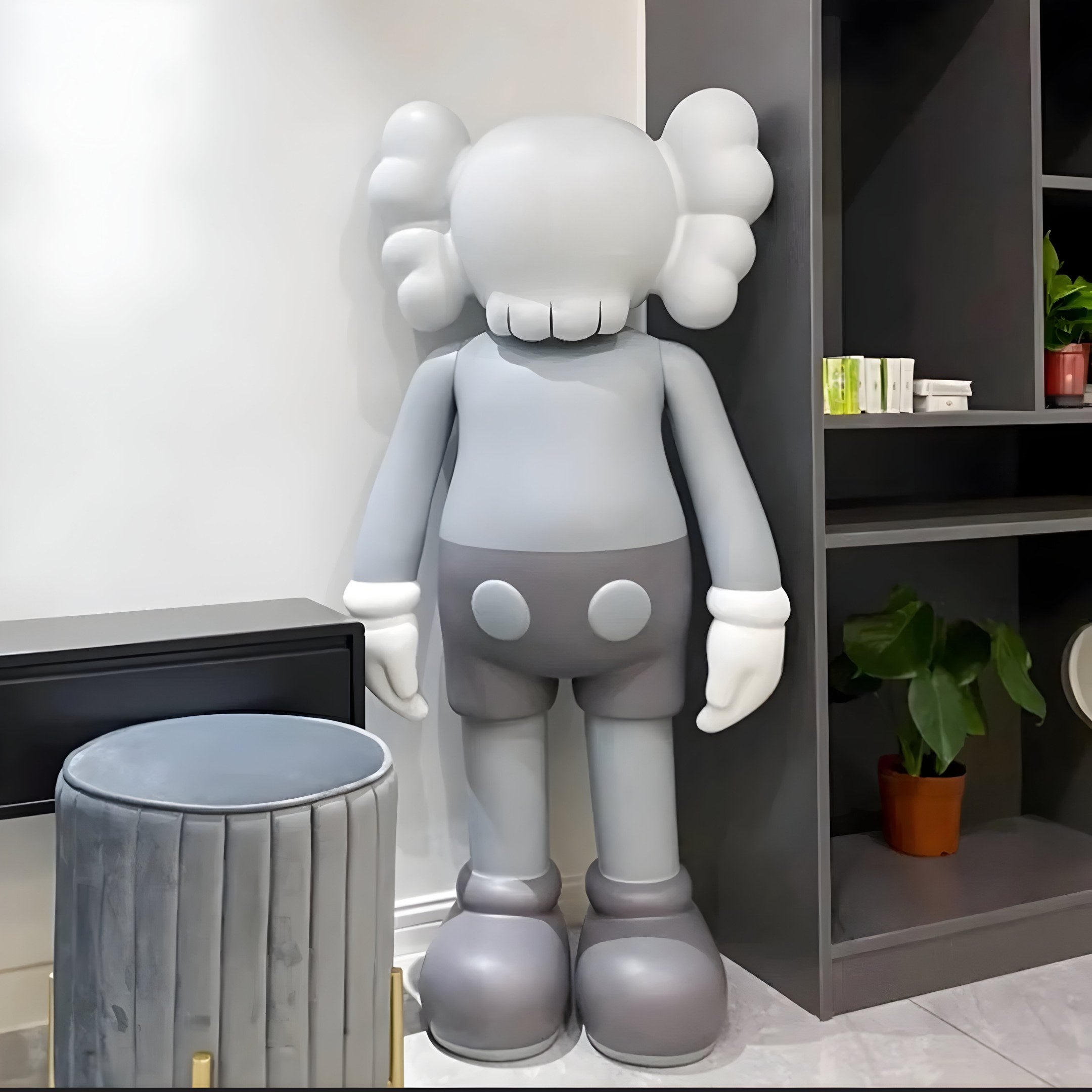 The Slay Grey Iconify Standing Sculpture - 100cm by Giant Sculptures stands in a room with its cartoonish big head, crossed-out eyes, and gloves. Positioned next to a black shelving unit with a plant and paired with a gray cylindrical stool, it adds contemporary flair to modern interiors.