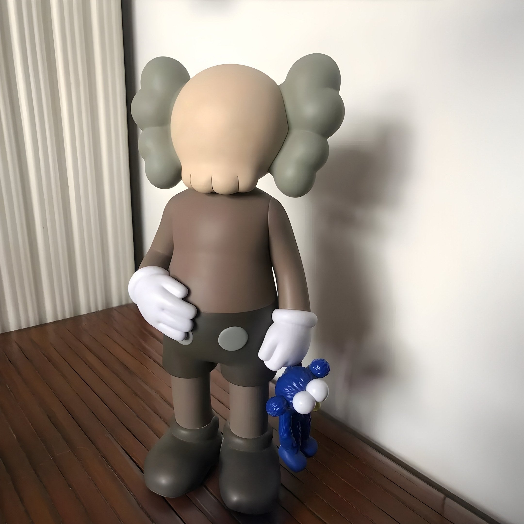 A 130cm Hazelnut Brown sculpture from Giant Sculptures with a cartoonish figure, round head, gray puffy ears, white gloves, and dark attire embodies contemporary aesthetics as it stands on a wooden surface holding an Iconify Blue Furry Toy Sculpture in its right hand.