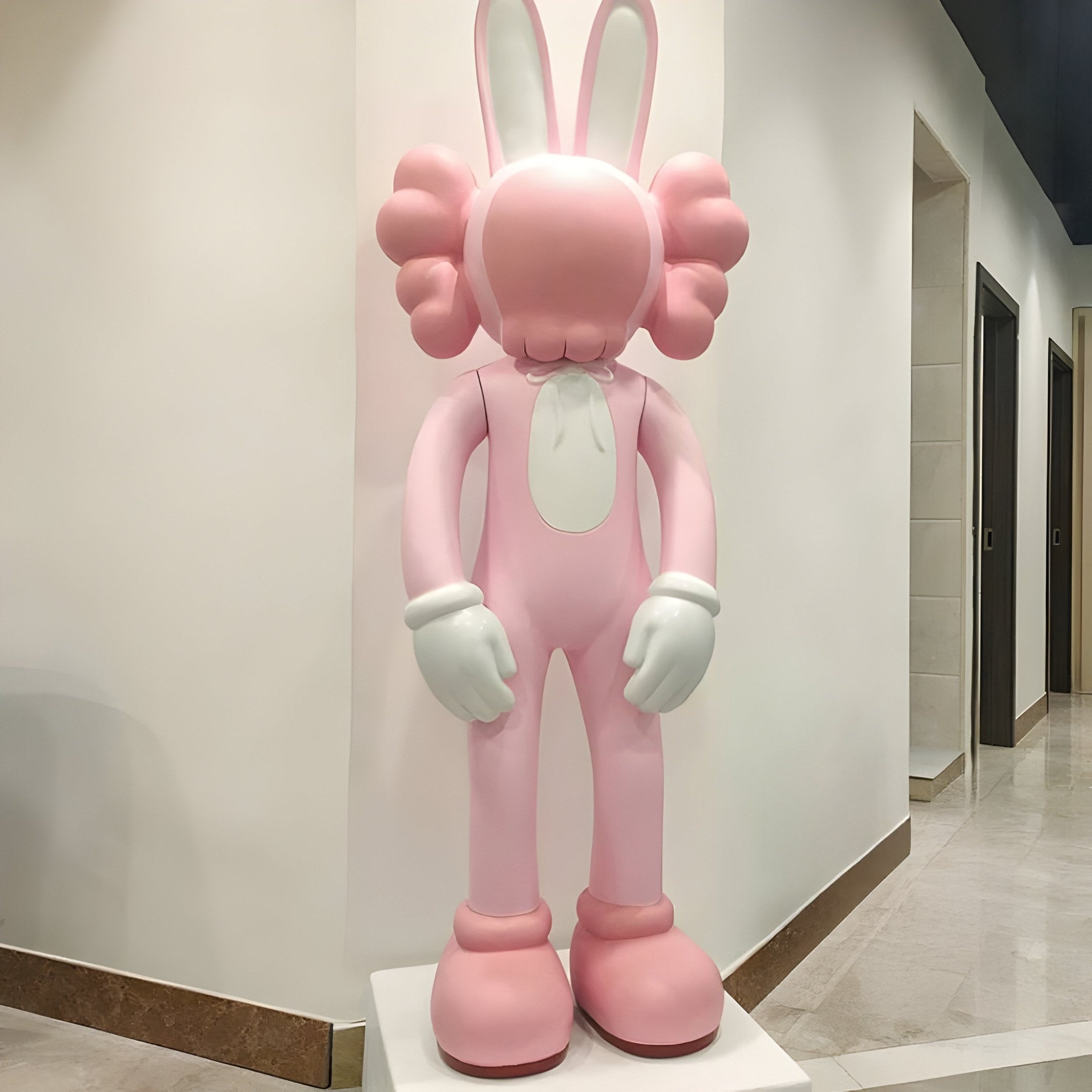 The Coral Pink Iconify Rabbit Sculpture by Giant Sculptures, standing 130cm tall with plush-like features and oversized gloves, rests on a white pedestal against a light wall. Its long ears and neutral expression epitomize contemporary design in the hallway.