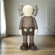 In a minimalist room with hazelnut brown floors, the Hazelnut Brown Iconify Standing Sculpture - 130cm by Giant Sculptures features a large figure with a round, featureless face, X eyes, padded ears, dark shorts with two dots, and shoes. White walls embrace a door and curtains for aesthetic appeal.