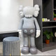 The Slay Grey Iconify Standing Sculpture by Giant Sculptures, measuring 130cm, stands in a corner alongside shelves and a gray stool. Its crossed-out eyes and gloved hands make it an artistic centerpiece against the rooms modern minimalistic design, complemented by a small plant on the shelf.