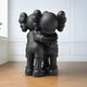 In a white room with wooden flooring, two Giant Sculptures Obsidian Black Iconify Embrace Sculptures (135cm) are embracing. These modern art pieces with oversized heads, X-shaped eyes, gloved hands, and ear-like protrusions create an emotional centerpiece.