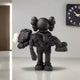 In a minimalist room, the Giant Sculptures Obsidian Black Iconify Rescuing Furry Sculpture (135cm) depicts a cartoonish figure with large ears gently cradling a smaller form with crossed-out eyes, while a modern clock adorns the wall.