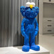 The Royal Blue Iconify Furry Sculpture by Giant Sculptures, standing 135cm tall, features a textured surface and large feet with big white eyes and a yellow nose. It stands upright in a modern room with gray and white tones.