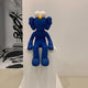 In a minimalist gallery, a 140cm Deep Blue Iconify Furry Sitting Sculpture by Giant Sculptures features white eyes and a yellow nose, perched on a ledge with its feet dangling. The contemporary pop art piece is highlighted by abstract black designs in the background.