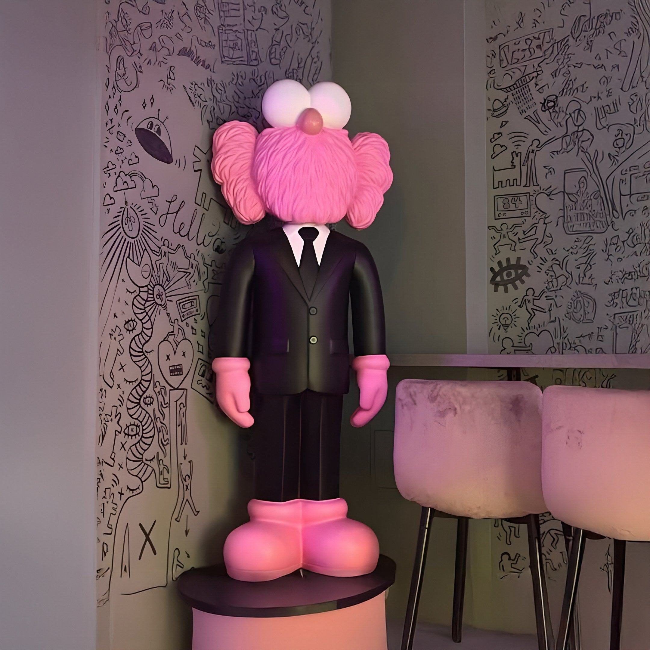 A pink cartoonish statue with exaggerated features, similar to the Black Suit Iconify CEO Sculpture by Giant Sculptures (147cm), stands on a pedestal. The background features doodle-like black wall art on a light surface, paired with tall stools in this contemporary design setting.