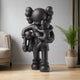 The Midnight Black Iconify Family Sculpture by Giant Sculptures is 200cm of modern design, depicting a figure with large gloves and a distinct head holding two smaller figures. Harmoniously complementing a cushioned chair and tall potted plant on wooden flooring, it symbolizes unity.