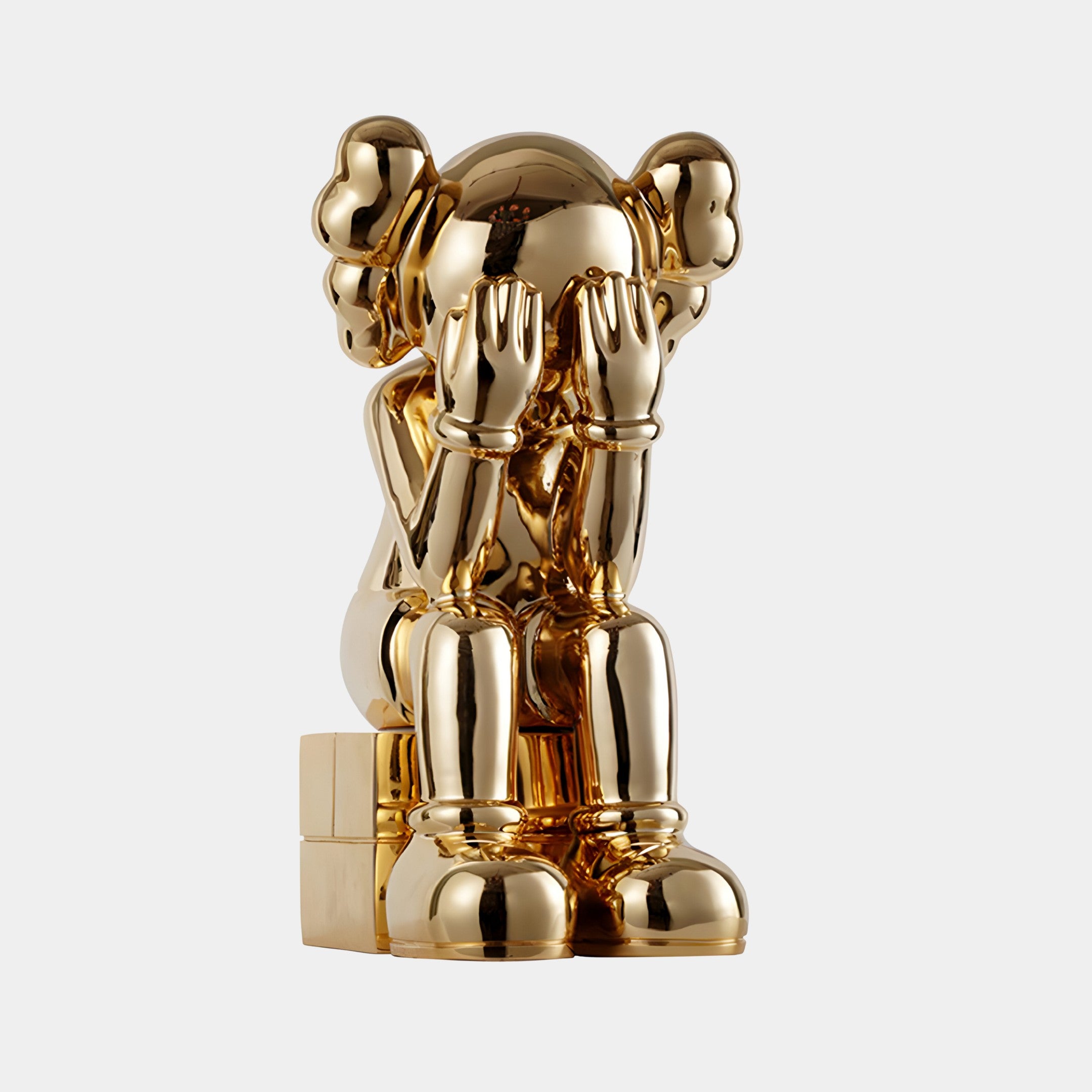 The Antique Gold Iconify Melancholy Sitting Sculpture by Giant Sculptures features a 70cm cartoon-style figure with exaggerated gloves and shoes sitting on a small block, covering its face with its hands and reflecting a sense of artistic melancholy.