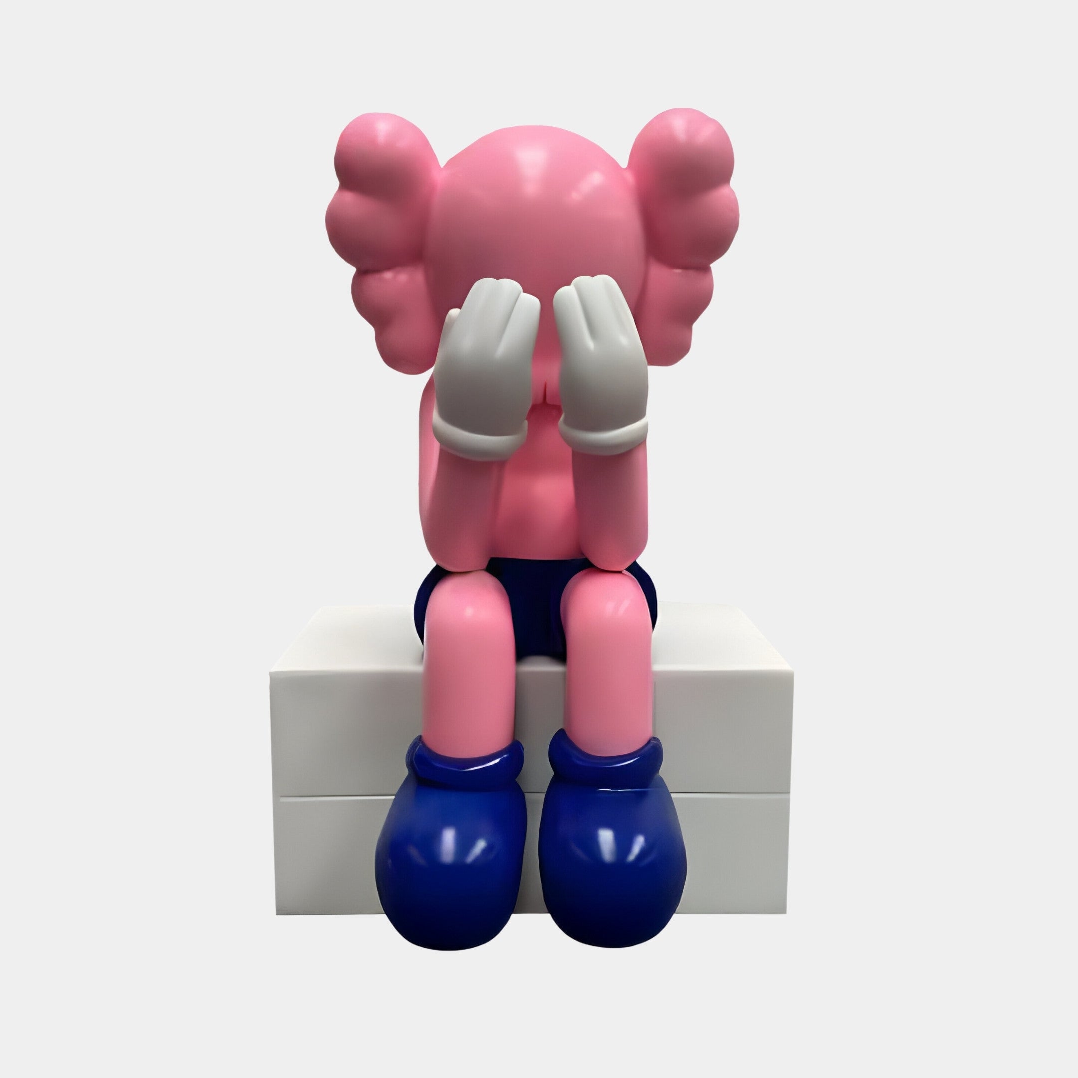 A Pedal Pink Iconify Melancholy Sitting Sculpture by Giant Sculptures sits on a white block in modern interiors. The 70cm resin figure, with gloved hands covering its face, resembles a child sporting blue shoes and rounded ear-like features in a sad posture.