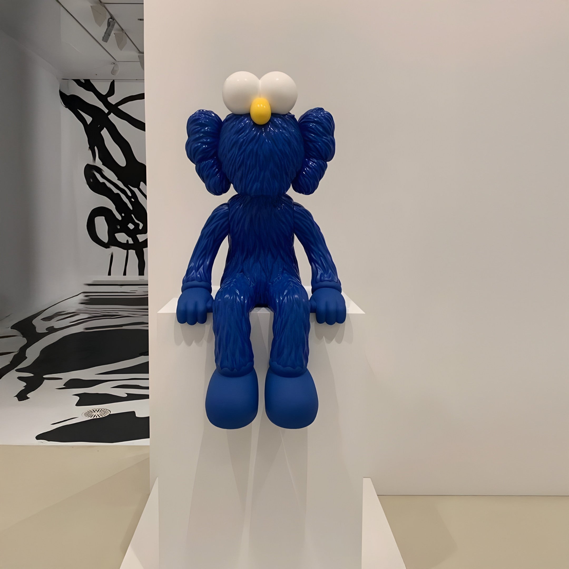 The Deep Blue Iconify Furry Sitting Sculpture - 70cm by Giant Sculptures, with its exaggerated, cartoonish features and large eyes and feet, rests on a white shelf in a gallery. The contemporary interiors enhance its artistic flair, framed by abstract black and white wall art.