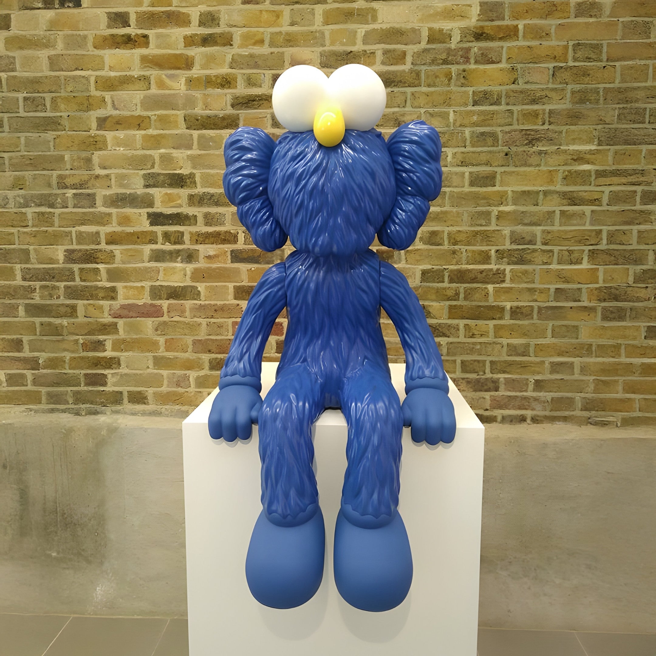 The Deep Blue Iconify Furry Sitting Sculpture - 70cm by Giant Sculptures, with its cartoon-like round eyes and yellow nose, sits artistically on a white pedestal. It stands out against the rustic brick wall, making it perfect for contemporary interiors.